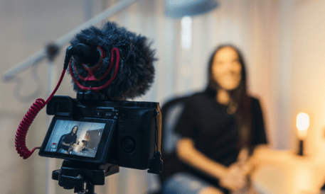 a professional camera and mic faces frames a women being interviewed with the photo focused on the camera as the women is blurred in the background